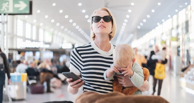 Mother traveling with child, holding his infant baby boy at airport terminal, checking flight schedule, waiting to board a plane. Travel with kids concept