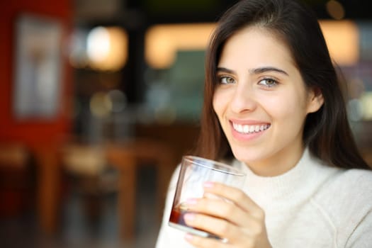 Happy woman holding soda glass in a bar looking at you