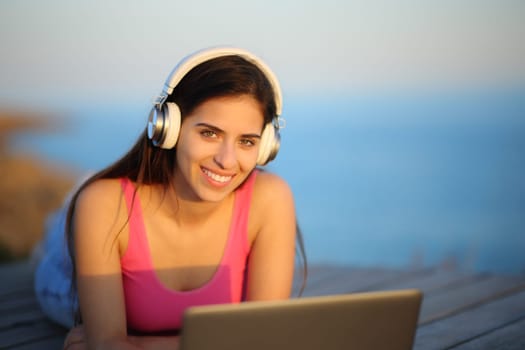 Happy woman with headphone and laptop posing looking at camera on the beach