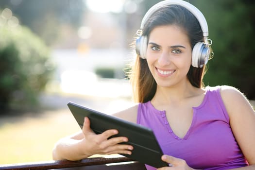 Happy woman with headphone and tablet looks at you sititng in a park
