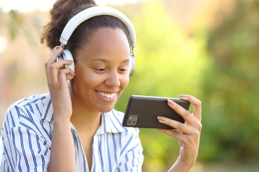 Happy black woman with headphone watching videos on phone in a park