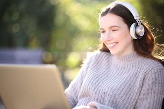 Happy woman listening and watching laptop content sitting in a park with headphone
