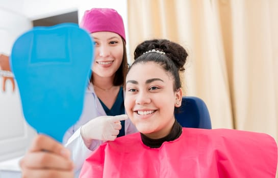 Satisfied patient in dental clinic looking at mirror, dentist next to satisfied patient smiling at dental mirror, female patient checking teeth after curing teeth in dental clinic.