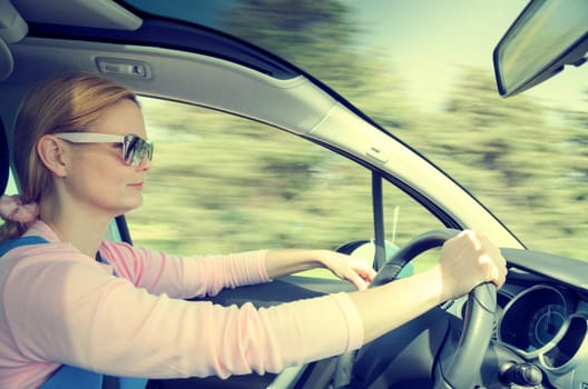Smiling beautiful woman in sunglasses driving at high speed car with panoramic windshield. Cross processing filtered and split toned internal stock photo with low shutter speed and blurred in motion natural background.