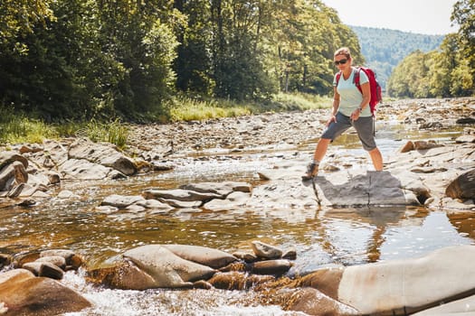 Trekking with backpack concept image. Female backpacker wearing trekking boots crossing mountain river. Woman hiking in mountains during summer trip. Vacation trip close to nature. Natural scenery