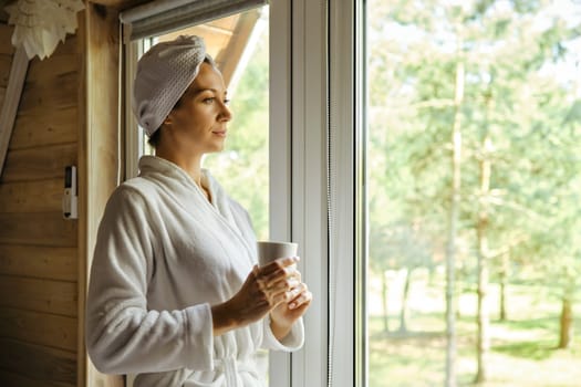 Peaceful woman in bathrobe came to the window of forest house after shower
