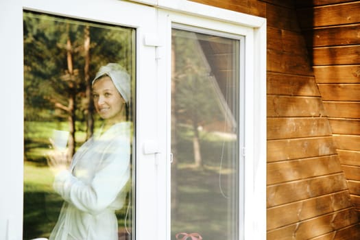Woman in bathrobe and towel on head leaned against doorway of wooden house and looks out