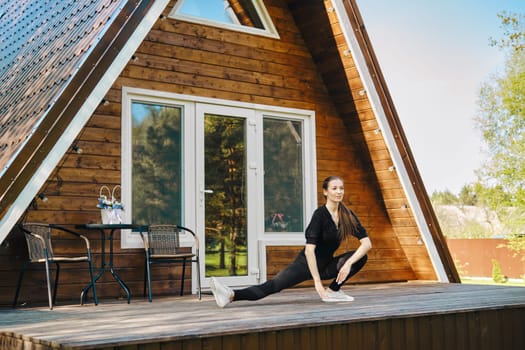 An adult woman in activewear does sports on terrace of her log cabin in sunny day