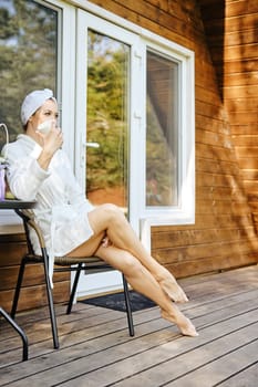 Thoughtful woman in bathrobe sitting in wicker chair on terrace of forest cabin