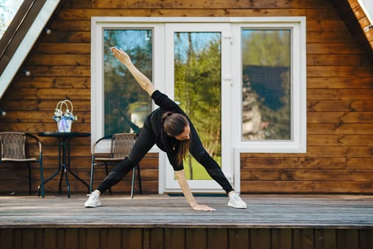 Athletic woman performs bends and spreads her arms to sides during morning workout on terrace of wooden house