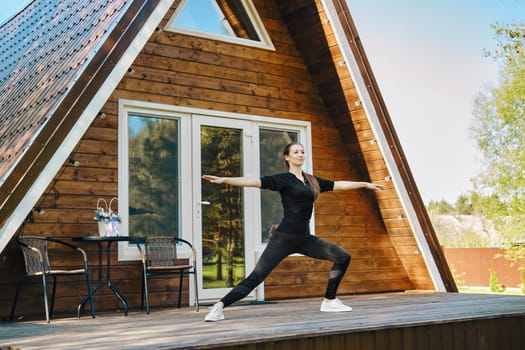 A flexible woman in sportswear performs stretching exercises on the terrace of a-frame house
