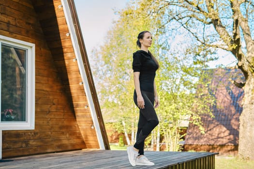 Concentrated woman in tracksuit went out on terrace of wooden house in morning and is preparing to do exercises