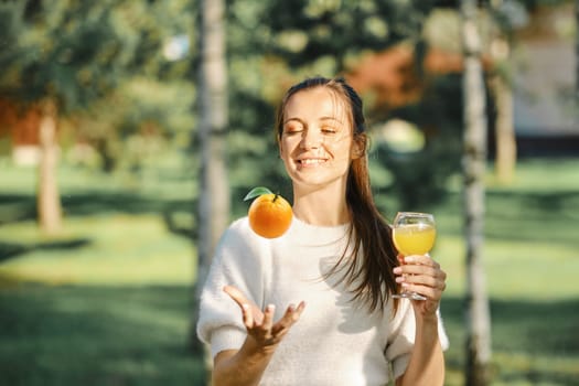 Joyful woman tossing orange with one hand while holding glass with fresh juice in another hand