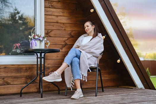 On warm evening, woman wraps herself in cozy blanket and rests on veranda of tiny house. Concept of small modern cabins for rest and escape to nature