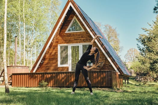 Active woman bends body to the sides during workout on lawn of her a-frame