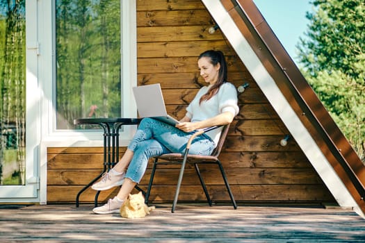 Young woman put laptop on her knees while resting on terrace of wooden a-frame cabin