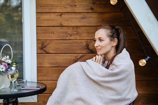 Dreamy cute woman sits on terrace wrapped in plaid with glass of wine