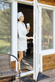 Pretty woman in bathrobe and cup of coffee in her hand goes out onto terrace of her forest house