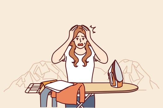 Housewife woman ironing clothes and clutching head in panic due to overload and fatigue. Housewife with iron stands near pile of clothes and needs assistant to take care of household