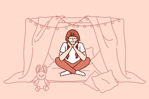 Little girl is sitting in home hut playing children games and dreaming of becoming princess with own castle. School-age girl smiles enjoying exciting game and cozy hut created from sheets and garlands