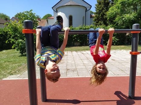 Sremska Mitrovica, Serbia, May 20, 2020. Children play and do sports on the playground. A girl and a boy of 6 and 7 years old hang upside down on gym bars