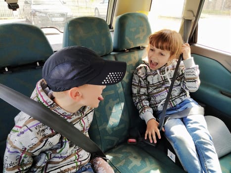 Children in the car. A boy and a girl are sitting in the back seat, laughing and grimacing. Children fasten their seat belts. A child in a cap. Children - twins of different faces dressed in the same clothes