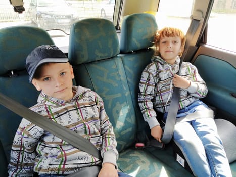 Children in the car. A boy and a girl are sitting in the back seat, laughing and grimacing. Children fasten their seat belts. A child in a cap. Children - twins of different faces dressed in the same clothes