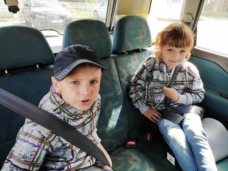 Children in the car. A boy and a girl are sitting in the back seat, laughing and grimacing. Children fasten their seat belts. A child in a cap. Children - twins of different faces dressed in the same clothes