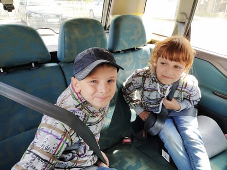 Children in the car. A boy and a girl are sitting in the back seat, laughing and grimacing. Children fasten their seat belts. A child in a cap. Children - twins of different faces dressed in the same clothes