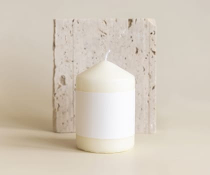 Candle with label on beige near cream travertine stone, Close up, copy space. Brand packaging mock up. Home decoration, minimal interior, Bohemian or scandinavian style