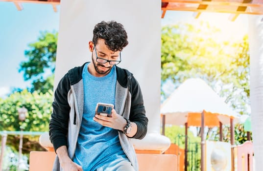 Smiling handsome man using a phone leaning on a wall in a park, Handsome guy in glasses leaning on a wall chatting on his cell phone, Teenage male leaning on a wall sending a text message