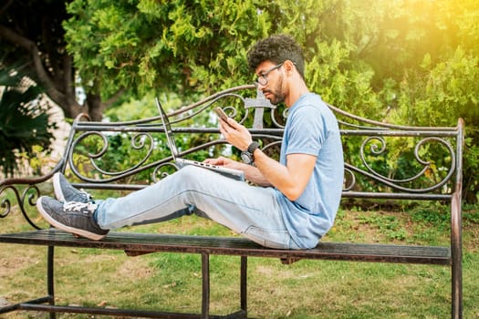Man in a park working online with laptop. Relaxed man working with laptop outdoor, Young man in a park with laptop and cell phone. Freelancer man sitting in a park using laptop and cellphone