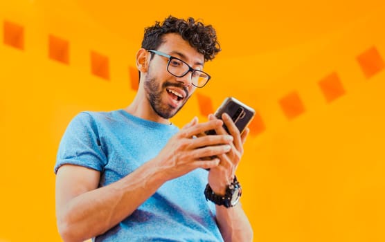 Happy guy sending a message with a cell phone near a yellow wall outdoors. Lifestyle of handsome bearded man using cell phone outdoors, Low angle view of young man using cell phone outdoors