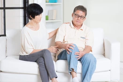 Asian old man knee pain, sitting on sofa with wife at home. Chinese family, senior retiree indoors living lifestyle.