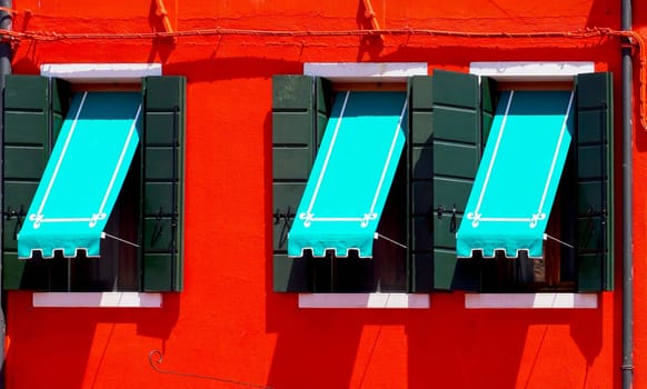 three Windows with blue canopy in Burano on red wall building architecture, Venice, Italy