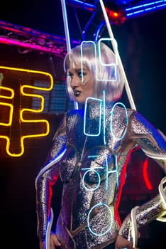 Blonde as a cyber robot. Girl in shining silver armor in neon. Blonde with fantastic makeup and image in dark key and colored light, posing in cyberspace. Letters are projected on her
