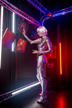 close-up of blonde in cyber-robot image. Girl in shining silver armor in neon, stands at full height and controls monitors. Blonde with fantastic makeup, poses in cyberspace. Monitors near her