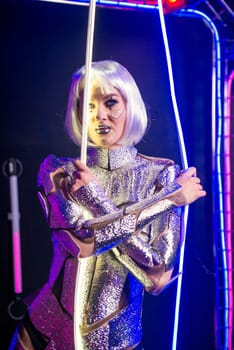 Blonde as a cyber robot. Girl in shining silver armor in neon. Blonde with fantastic makeup and image in dark key and colored light, posing in cyberspace. In her hands she holds wires