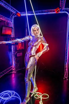 Blonde as a cyber robot. Girl in shining silver armor in neon. Blonde with fantastic makeup and image in dark key and colored light, posing in cyberspace. Around her hands and body wires, neon color