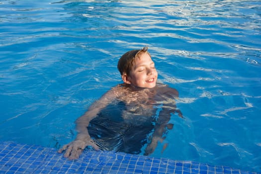 A boy holds on to the edge of a swimming pool and laughs as he looks into the sun