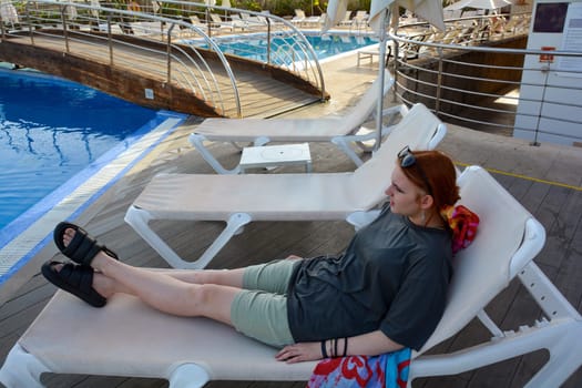 A young teenage girl, dressed in clothes, lies on a sun lounger by the swimming pool