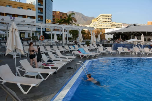 A boy and a girl at the hotel pool with blue water and some sun loungers and with mountains in the background