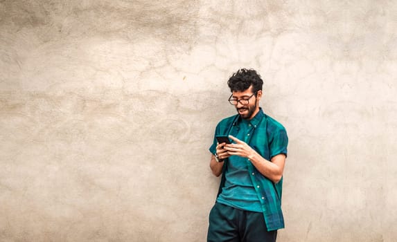 Happy handsome guy leaning against the wall using his cell phone, happy latin man with his cell phone leaning against a wall with copy space, Person smiling phone with space for text