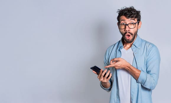 Amazed handsome man pointing at his cell phone isolated, surprised person pointing at his cell phone isolated. Isolated teenager surprised of a promotion on his cell phone