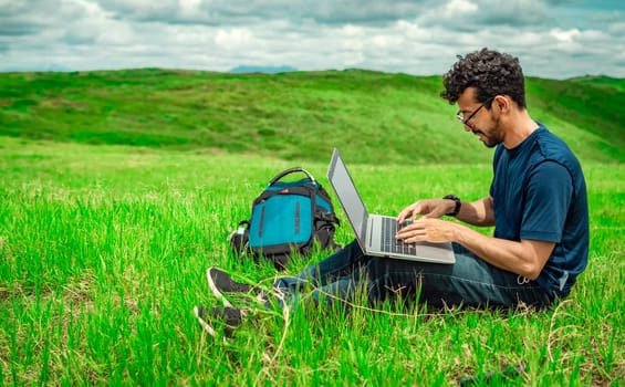 Close up of man sitting on green grass working with his laptop. Smiling freelancer guy working from the field. Smiling man with laptop sitting on the grass in the field