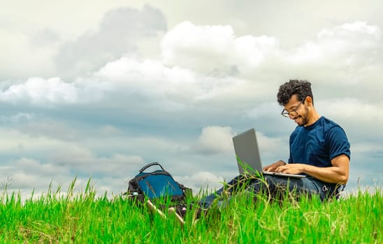 Smiling freelancer man sitting with laptop and backpack in field. Portrait of a man sitting on the grass of the field working with his laptop. Freelancer man concept working from the field
