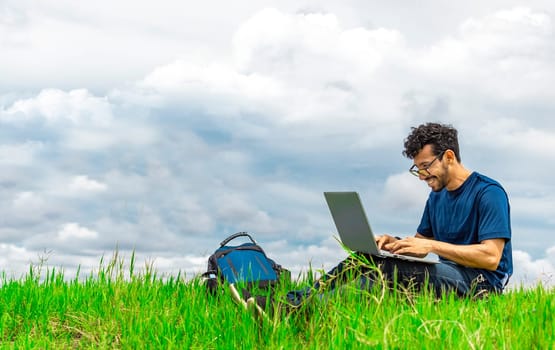 Smiling man with laptop sitting on the grass in the field. Side view of man sitting on green grass working with his laptop. Smiling freelancer guy working from the field