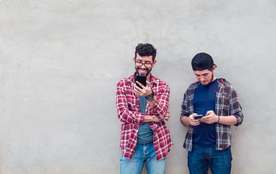 Two smiling friends leaning on a wall checking their cell phones, Friends leaning on a wall texting on their phones. Friend showing cell phone to his friend, Smiling friends checking cell phones