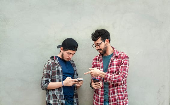 Two young friends leaning against a wall checking their cell phones, Two friends leaning against a wall looking at the content on their phones. Friend showing cell phone to his friend outdoors