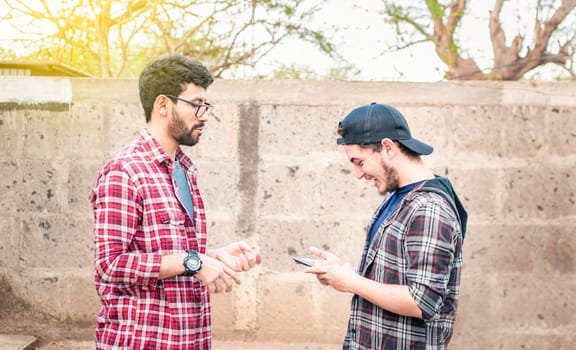 Side view of two young friends chatting outdoors, two friends chatting while smiling and using cellphone. View of two teenagers talking face to face while using the cell phone